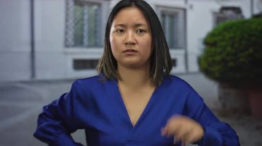 Mavi gömlekli genç bir kadın, şehir geçmişiyle çerçevelenmiş belirsiz bir ifadeyle şehir caddesinde el kol hareketi yapıyor, gündüz vakti dışarıdaki merakları aktarıyor..