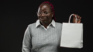 Kırmızı örgülü gözlüklü bir kadın siyah arka planda beyaz bir alışveriş çantası tutuyor, modern ve soyutlanmış bir tarzı ve tarzı var..