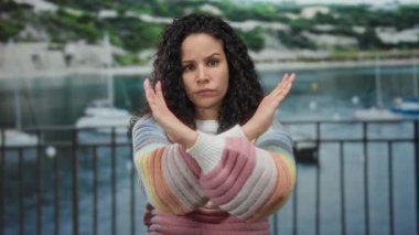 Renkli süveter giyen kadın, deniz kıyısında el hareketleriyle arka planda tekneleri olan, açık hava ortamında reddeden ya da anlaşmazlık yaşayan kadınları reddediyor..