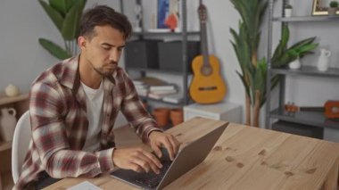 Modern bir ofiste dizüstü bilgisayarda çalışan İspanyol bir adam, arka planda bitkiler ve gitarla bir iç mekanda stres gösteriyor, yoğun bir iş ortamını yansıtıyor..