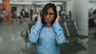 Mavi gömlekli kadın yoğun havaalanı terminalinde mizahi bir şekilde poz veriyor. Arka planı bulanık. Kalabalık, kapalı bir ortamda neşeli bir seyahat havası taşıyor..