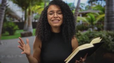 Gözlüklü genç bir kadın, tropikal ağaçlarla çevrili yemyeşil bir park ortamında tutkulu bir şekilde kitap okuyor, dışarıda eğitici ve ilham verici bir atmosfer yaratıyor..