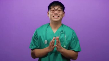 Yeşil önlüklü genç adam, izole edilmiş mor bir arka plana gülümsüyor ve steteskop boynunda neşeli bir sağlık hizmeti sunuyor.
