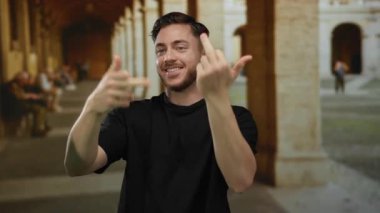 Sakallı genç bir adam eski bir üniversite koridorunda el kol hareketleri yaparak modern ifade ve klasik mimari arasındaki zıtlığı yakalıyor..