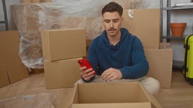 Genç bir adam yeni evinde kutuları yerleştirirken akıllı telefonunu kontrol ediyor. Etrafı karton kutularla çevrili. Parlak bir oturma odasında..