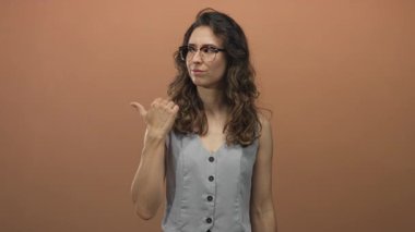 Kolsuz yelek ve gözlüklü kadın kameraya kaşlarını çatarken stüdyoda başparmağıyla işaret ediyor..
