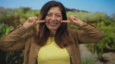 Kadın geniş gülümseme ile yeşil yapraklar ile güneşli orman sahnesinde iki işaret parmağı ile yanaklarını çimdikler;.