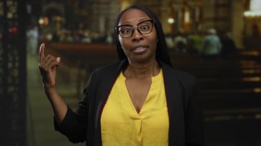 Sarı tişörtlü ve gözlüklü kadın şehir sokaklarında duygularını ifade ediyor ve arka planda insanlar var..