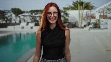 Lüks bir otel havuzunun yanında neşeli bir şekilde gülümseyen bir kadın açık hava tatil köyü ortamında kızıl saçlar, gözlükler ve modern ve ferahlatıcı bir havası olan günlük kıyafetler sergiliyor..