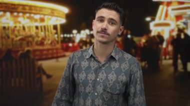 Genç İspanyol adam gece fuarında arka planda atlıkarınca ışıklarıyla eğleniyor, desenli gömlek giyiyor ve canlı park ortamında düşünceli bir şekilde poz veriyor..
