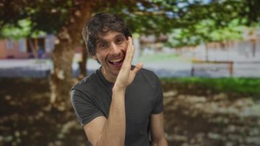 Gri tişörtlü adam ormanda güneş ışığında yeşil yaprakların altında kıt kanaat ötüyor; neşeli eğlence enerjisi.