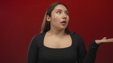 Avuç içleri kalkık, siyah, uzun kollu bir bluz giyen İspanyol genç bir kadın ve kırmızı bir stüdyo duvarına bakan gözleri var..