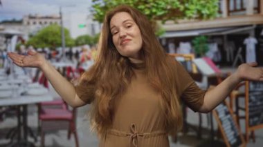 Kadın, kahverengi elbise ve omuzlarından dökülen gevşek saçlarla sokak restoranının terasında çıplak kollarını silker; belirsizlik.