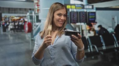 Havaalanı terminalinde elinde ahşap çatal ve plastik yiyecek konteynırını tutan kadın gülümsüyor..