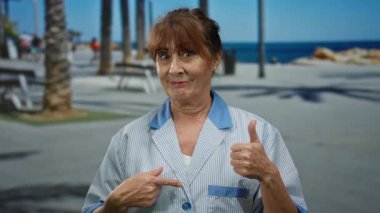 Kıdemli İspanyol kadın temizlikçi arka planda palmiye ağaçları olan deniz kenarındaki gezinti güvertesine başparmak işareti veriyor. Üniforma giyiyor ve gülümsüyor. Açık hava ortamında pozitiflik ve güven gösterisi yapıyor..