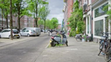Bulanık ağaçlar, park etmiş arabalar, bisikletler ve kaldırımda kuyruk oluşturan scooterlarla dolu Amsterdam caddesinin odaklanmamış görüntüsü, Hollanda 'da tipik bir şehir manzarası yaratıyor..