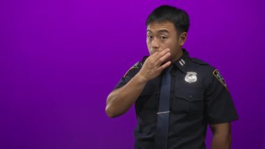 Polis üniformalı ve rozetli bir adam mor stüdyoda sessiz kalmak için parmaklarını dudaklarına uzatıyor..