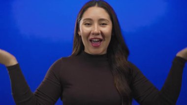 Latin kökenli genç bir kadın, parlak mavi stüdyo oturumunda el ele tutuşup gülümsüyor..