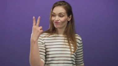 Çizgili gömlek giyen bir kadın mor bir stüdyoda parmaklarıyla hashtag jesti oluşturuyor; neşeli bir bağlantı.