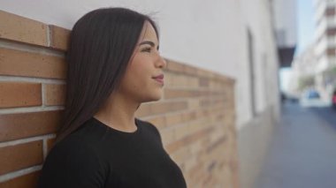 Uzun saçlı genç bir kadın şehir sokaklarında duvara yaslanıyor, sakin bir ifade sergiliyor ve açık havada düşünceli davranışlar sergiliyor..
