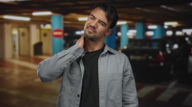 Kapalı otoparktaki genç İspanyol bir adam park halindeki bir arabanın yanında rahatsız bir şekilde boynuna masaj yapıyor.
