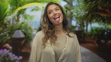 Bej gömlekli kadın, yemyeşil çiçeklerle ve çiçek açan çiçeklerle çevrili bir orman geçidinde dilini dışarı çıkarıyor..