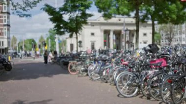 Amsterdam, Hollanda 'da, güneşli bir günün tadını çıkaran yayalar tarafından çevrelenmiş, dikkati dağılmış şehir manzaralı cadde boyunca park etmiş bisikletlerin odak dışı görüntüsü..
