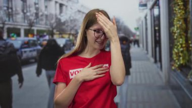 Kırmızı gönüllü gömleği giyen kadın kalabalık şehir caddesinde alnına ve göğsüne dokunuyor..