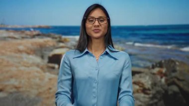 Kumsalda gülümseyen, parmaklarıyla kalp hareketleri yapan, gözlük takan, mavi gömlekli, arka planda deniz olan güneşli bir deniz gününün tadını çıkaran bir kadın.