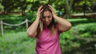 Huzurlu bir park ortamında hayal kırıklığını ifade eden, pembe bir gömlek giyen ve açık havada yemyeşil bir yerde dikilirken elleriyle duygularını gösteren bir kadın..