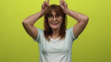 Gözlüklü yaşlı İspanyol kadın, parlak sarı bir arka plana karşı şakacı poz veriyor. Elleri boynuz şeklinde sıcacık gülümsüyor..