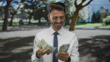 Beyaz gömlekli genç İspanyol adam orman açıklığında elleri olan dirhem banknotları sayıyor; refah.