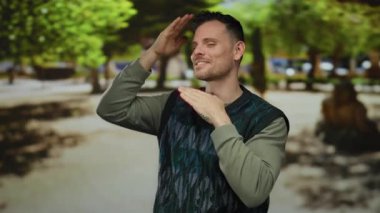 Genç adam arka planda ağaçlarla güneşli bir parkta neşeli poz veriyor, neşe ve karizmayı ifade ediyor, sıradan bir süveter giyiyor, gülümsüyor, elleri çerçeveli bir jest yapıyor..