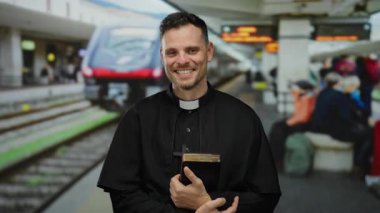 Tren istasyonunda elinde İncil 'le gezen rahip, gün içinde arka planda insanlarla gülümsüyor.