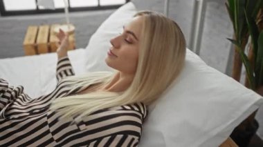 Çizgili gömlekli sarışın kadın, eli yastıkta, gözleri kapalı olarak yatakta uzanıyor. Stüdyoda beyaz yatağın arasında, huzur içinde..