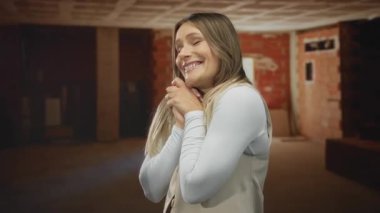 Sarışın bir kadın, duvarları ve kirişleri açık bir inşaat alanında heyecanlı bir şekilde gülümsüyor. Gelişmekte olan bir yerde mutluluk ve beklenti simgeliyor..