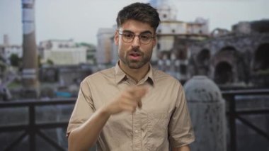 İspanyol gözlüklü ve bej gömlekli adam antik Roma harabelerinin önünde telefon jesti yapıyor..