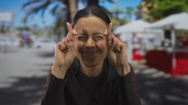 Latin kökenli genç bir kadın gün ışığının altında, arka planda bulanık palmiye ağaçları ve kafelerle şehir ortamında umut ya da batıl inanç olduğunu düşündürüyor.