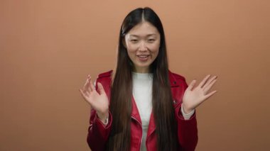 Kırmızı ceketli bir kadın, izole edilmiş kahverengi bir arka plana karşı sürpriz bir hareketle gülümsüyor. Duygularını ifade ediyor..