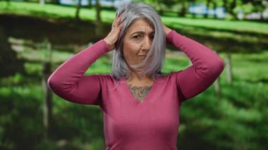 Gri saçlı, canlı bir açık hava parkında poz veren, değişik duygular ve jestler sergileyen, pembe bir kazak ve dövme ile vurgulanmış, kişiliği ve tarzı olan yaşlı bir kadın..