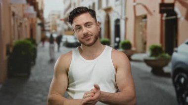 Şehir sokağında sakallı genç bir adam. Üzerinde beyaz bir atlet var. Şehrin arka planında binalar ve arabalar var..