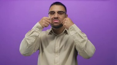 Bej gömlek giyen İspanyol genç adam mor stüdyo setinde sahte ağlama hareketi ile gözlerini ovuşturur, alaycılık alay eder..