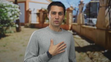 Genç adam bir park ortamında ayakta duruyor, elini göğsüne koyup düşüncelere dalıyor, ağaçların arkasına ve güneşli bir güne karşı..