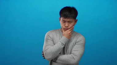 Genç bir adam canlı mavi bir duvar üzerinde kafa yoruyor, izole edilmiş bir stüdyoda düşünceli bir ifade sergiliyor ve düşünüyor..