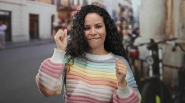 Şehir sokaklarında gülümseyen bir kadın renkli bir kazak giyiyor açık şehir ortamındaki kıvırcık saçlarıyla neşe saçıyor ve mutluluğunu ve hayat dolu tarzını vurguluyor..