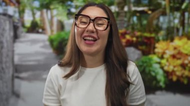 Gözlüklü kadın ağaçlarla çevrili yeşil bir parkta güneş ışığı altında gülümsüyor..