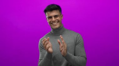 Genç adam neşe ve karizma saçan canlı pembe arka plana karşı gözlük ve kazak içinde mutlu bir şekilde alkışlıyor.