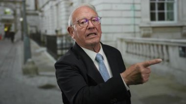 Üst düzey bir adam takım elbise giyip açık bir sokak ortamını işaret ediyor. Bu da tarihi binaların arka planına karşı hayal kırıklığını ya da vurguyu gösteriyor..