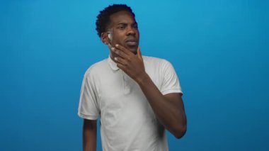 Beyaz gömleğin üzerinde çeneye dokunan bir adam stüdyoda kameradan dışarı bakıyor; düşünceli iç gözlem..