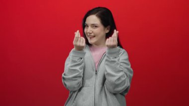 Gri kapüşonlu bir kadın kırmızı arka plana karşı para hareketi yaparak neşeli bir güven gösterisi yapıyor..